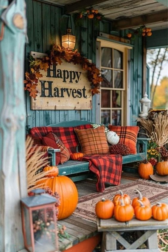 This charming porch is the perfect autumn retreat, radiating cozy and festive vibes. The rustic wooden bench is piled high with plaid pillows and a matching blanket in rich fall hues of red and orange, creating a warm and inviting spot to sit and enjoy the season. Above the bench, a 'Happy Harvest' sign, framed by a garland of autumn leaves, adds a festive touch to the space. The porch is adorned with an array of pumpkins, from the large one at the foot of the bench to the smaller ones artfully arranged on the table. A lantern filled with seasonal accents and a bundle of wheat in a vase complete the look, making this porch the ultimate spot to celebrate the harvest season.