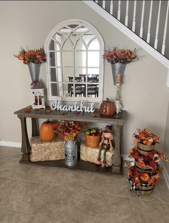 This entryway is a delightful tribute to the spirit of fall and thanksgiving! The console table is beautifully decorated with a mix of rustic and seasonal elements. A large mirror with an arched frame adds depth and elegance, while galvanized metal vases filled with vibrant autumn florals bring color to the space. The word 'Thankful' takes center stage on the table, surrounded by pumpkins, lanterns, and a charming scarecrow. Below, bales of hay serve as a base for more fall decor, including a classic orange pumpkin and a basket of apples, evoking the harvest season. To the side, stacked bushel baskets overflowing with leaves and mini pumpkins complete this cozy, welcoming setup, making it the perfect spot to greet guests with a warm fall vibe.