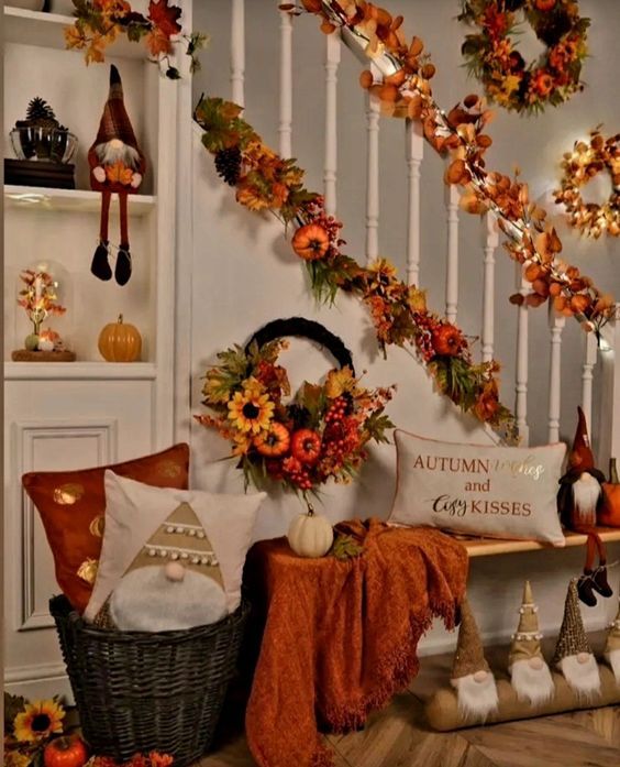 This staircase area has been transformed into a cozy and whimsical fall retreat! The banister is wrapped in a gorgeous garland of autumn leaves and flowers, creating a cascading effect that brings warmth and color to the space. Below, a soft, burnt orange throw drapes over a small table adorned with a festive wreath and a cute pumpkin, adding layers of texture. The corner is made even more inviting with pillows, including one with a charming gnome and another that reads 'Autumn Leaves and Cozy Kisses,' setting a cozy tone. A basket filled with more pillows and a trio of adorable gnome figurines bring a touch of playfulness to the decor. The shelf on the wall adds extra seasonal charm with a small pumpkin and twinkling lights, making this space feel like the perfect nook to enjoy all the best parts of autumn.