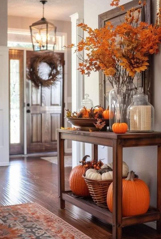 This entryway is a picture-perfect example of fall decor at its finest! A wooden console table greets you with a beautiful display of vibrant orange leaves arranged in a tall vase, creating a striking focal point. The table is adorned with small pumpkins, glass cloches, and a mix of seasonal elements that bring warmth and charm to the space. Below, a wicker basket filled with white pumpkins and additional large orange pumpkins add a grounded, earthy touch. The open front door, with a simple yet elegant wreath, invites you into this cozy, welcoming home, where the autumn vibes are in full swing. The rich tones of the hardwood floor and the patterned rug enhance the overall warmth, making this entryway the perfect introduction to the fall season.