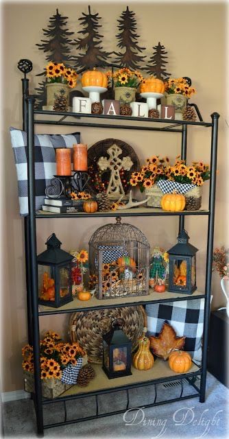 This shelf is a beautifully curated fall display, bursting with charm and seasonal flair! Each level is adorned with a variety of autumnal decorations, creating a layered and inviting look. The top shelf features white pumpkins, rustic candles, and blocks spelling out 'FALL,' all framed by a backdrop of metal pine trees that add a woodland touch. Buffalo check pillows and fabric accents throughout the display bring a cozy, farmhouse vibe, while sunflowers add pops of bright, cheerful color. Lanterns filled with tiny pumpkins and fall leaves, along with a decorative birdcage, add a rustic elegance to the arrangement. The bottom shelf ties it all together with more seasonal elements, including a woven basket and additional sunflowers, creating a warm and welcoming space perfect for the autumn season.