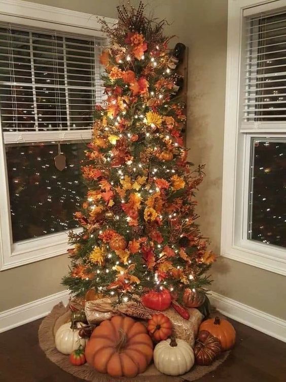 Adorned with vibrant orange, red, and yellow leaves, as well as autumn flowers and pinecones, this tree perfectly captures the essence of the season. Twinkling white lights are woven throughout, adding a warm and magical glow to the arrangement. At the base, a variety of pumpkins in different sizes and colors, from deep orange to creamy white, create a harvest-inspired foundation that ties the whole look together. This unique blend of fall and festive elements makes this tree a stunning centerpiece for celebrating both autumn and the upcoming holiday season.