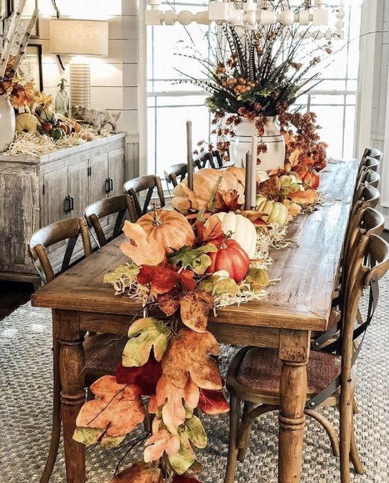 This dining room is the epitome of fall festivity, with its stunning centerpiece that runs the length of the rustic wooden table. A vibrant garland of oversized leaves, pumpkins, and gourds winds its way down the middle, creating a rich tapestry of autumn colors. Tall candlesticks add a touch of elegance, while the large vase filled with seasonal foliage brings height and drama to the setting. The sideboard echoes the theme with its own arrangement of pumpkins and natural elements, making this room a perfect spot for a cozy fall dinner with family and friends.