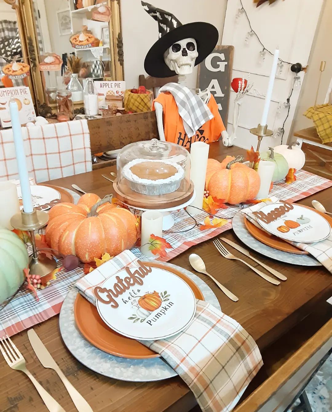 This festive dining table is ready for a fun and spooky fall gathering! The centerpiece of the table is a whimsical skeleton, dressed in an orange shirt and a witch's hat, adding a playful touch to the seasonal decor. The table is set with plaid napkins and 'Grateful Hello Pumpkin' plates, perfectly matching the fall theme. Glittering pumpkins and scattered autumn leaves add color and sparkle, while tall candles in brass holders bring warmth and elegance to the setup. A delicious-looking pie under a glass cloche takes center stage, making this table as tasty as it is festive. With its mix of cozy and spooky elements, this table is perfect for celebrating the harvest season with a hint of Halloween fun!