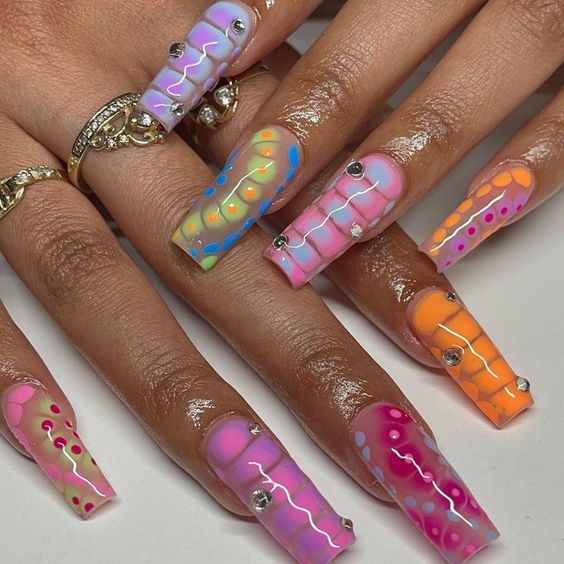 This nail art design features a playful blend of pastel colors in a snake skin pattern, accentuated with rhinestones for added sparkle. Each nail showcases a unique combination of pink, orange, blue, and yellow hues, creating a fun and whimsical look that’s perfect for summer.