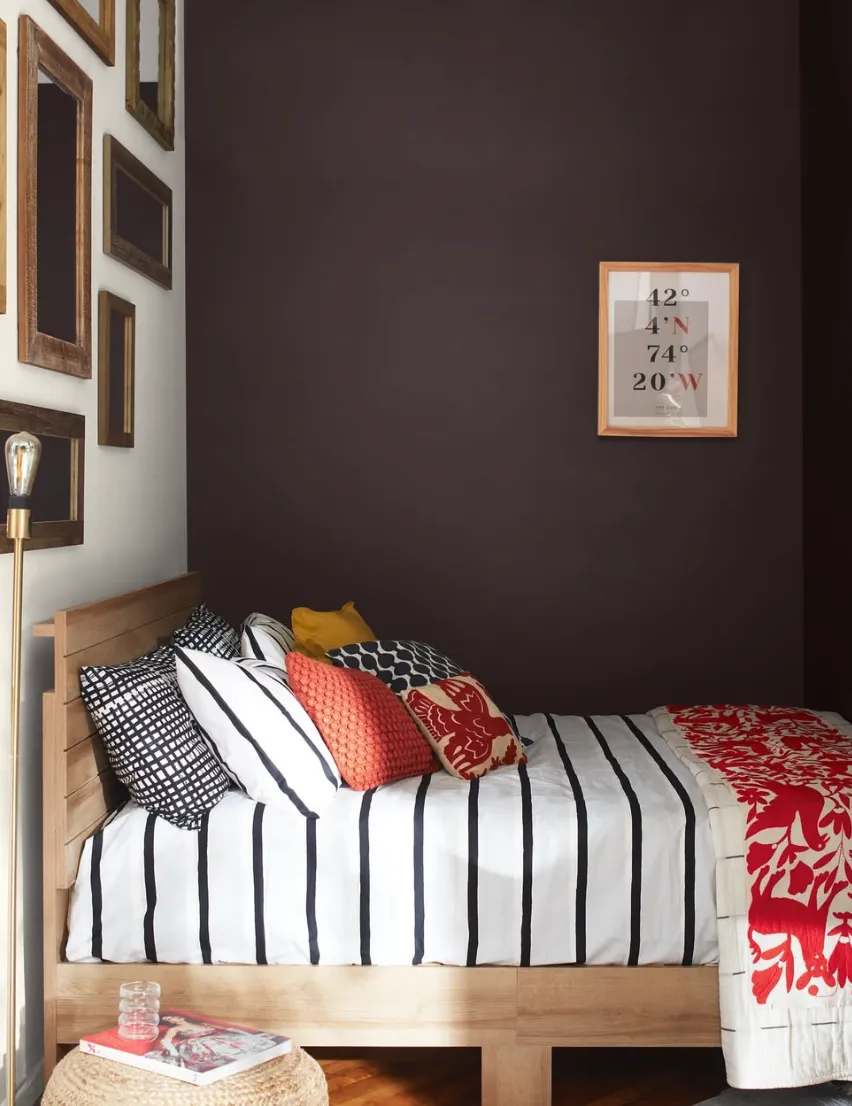 Dark and Moody EleganceA stunning bedroom with a deep chocolate brown statement wall. The rich, dark color adds a cozy and dramatic atmosphere to the space. To contrast the bold wall, light-colored bedding with crisp black and white stripes is paired with vibrant pops of red, yellow, and patterned cushions, creating a playful yet sophisticated look. The addition of a gallery wall of eclectic frames enhances the depth and texture of the design.