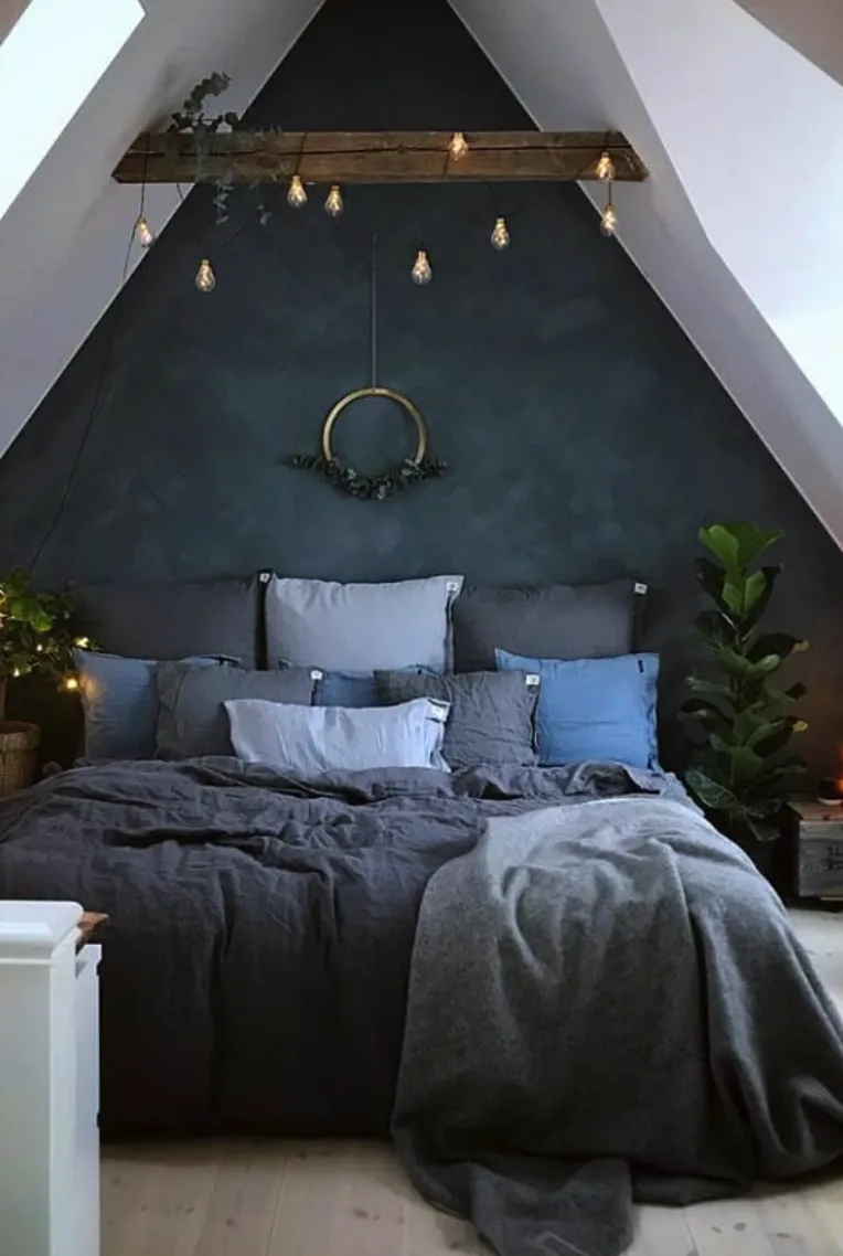 Moody EleganceA deep charcoal accent wall paired with moody lighting transforms this attic into a dramatic yet relaxing space. Cozy layers of blue and gray bedding invite you to unwind, while string lights and greenery add a magical touch.