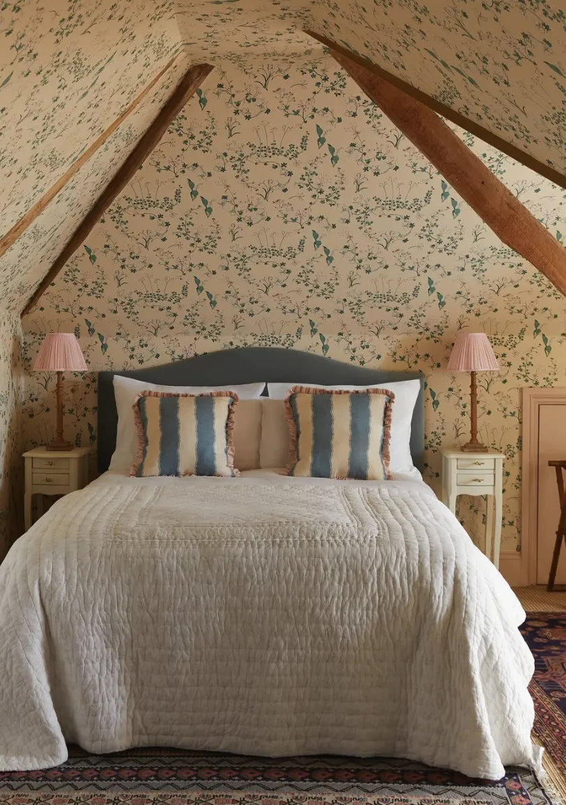 Floral Cozy HavenThis floral-themed bedroom takes full advantage of the attic’s sloped ceiling with a wallpapered accent wall. The textured quilt and soft lighting from pink bedside lamps enhance the inviting atmosphere.