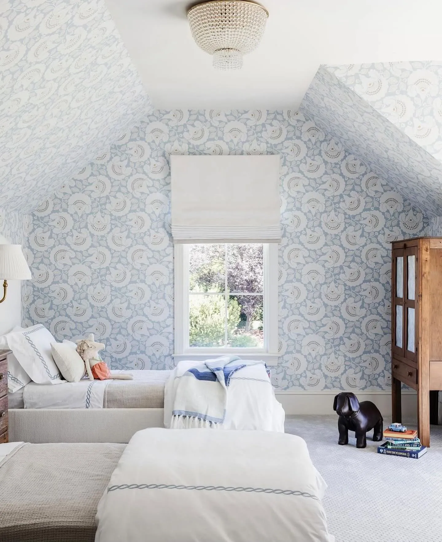 Elegant SymmetryTwin beds under a sloped ceiling make this attic bedroom both functional and beautiful. The soft blue wallpaper and coordinated bedding bring a cohesive and calming look, while the central window maximizes natural light for a fresh and airy feel.