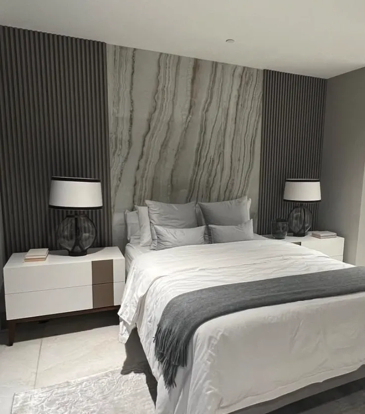 This bedroom features a striking tile accent wall behind the bed, with vertical, textured stone-like tiles in muted neutral tones. The natural textures add depth and sophistication to the space, perfectly balanced by minimalistic white side tables and elegant black-and-white lamps. The design exudes modern elegance and can be achieved by using lightweight, peel-and-stick tile panels for a DIY-friendly approach or opting for real stone tiles with professional installation.