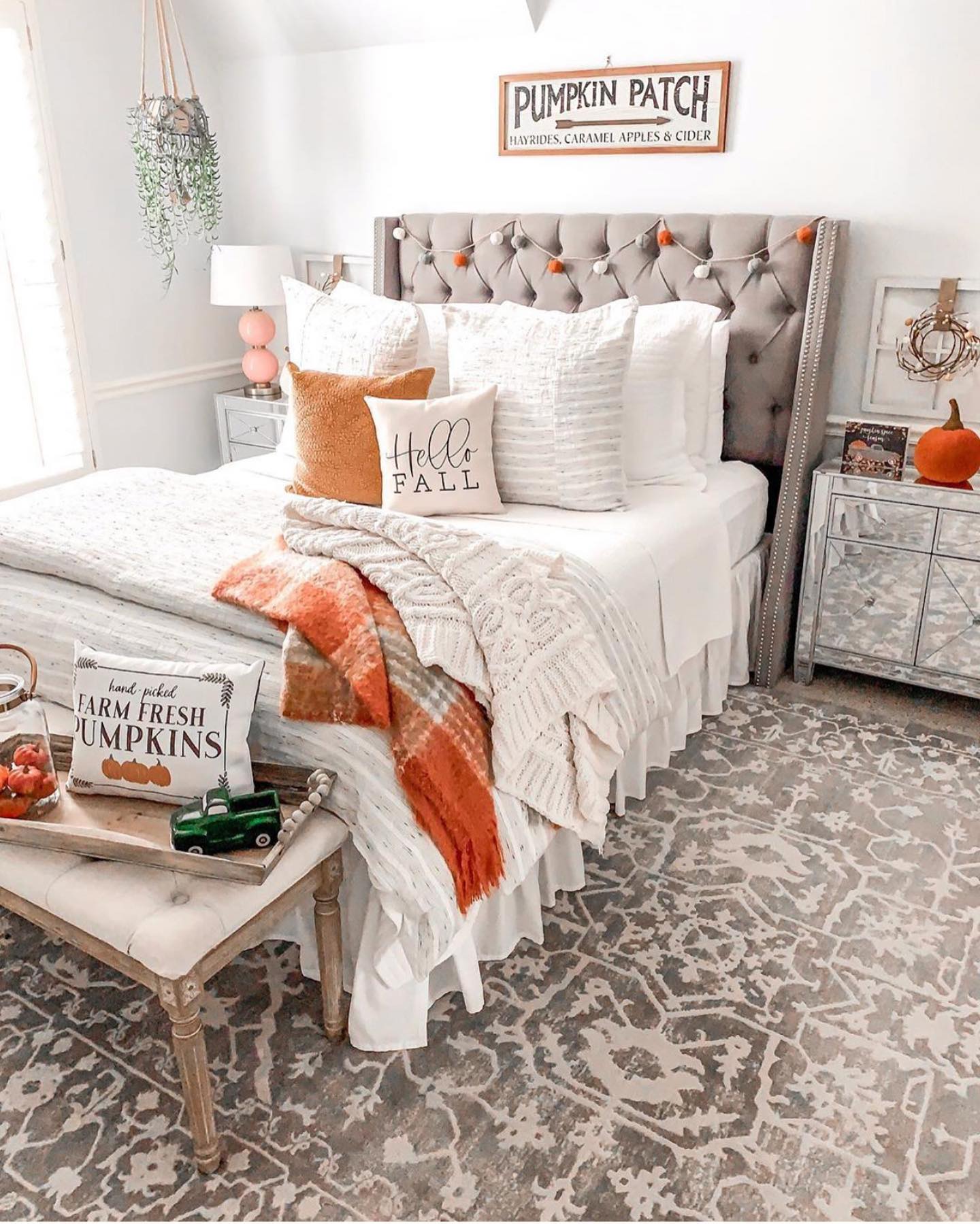 This farmhouse-inspired bedroom is ready to welcome fall with open arms! The neutral tones of the room are beautifully accented with autumnal touches that add warmth and coziness. The tufted headboard is adorned with a charming pom-pom garland, while the bed is layered with plush pillows, including a standout 'Hello Fall' cushion that sets the seasonal tone. A cozy plaid blanket draped at the foot of the bed adds a pop of color and texture. The 'Pumpkin Patch' sign above the bed brings a rustic charm, perfectly complemented by the small pumpkin and wreath on the bedside table. The bench at the end of the bed is styled with a 'Farm Fresh Pumpkins' pillow and a decorative tray, adding to the room's inviting fall vibe. This bedroom is the perfect blend of farmhouse style and seasonal decor, making it a warm and welcoming retreat for the autumn season.