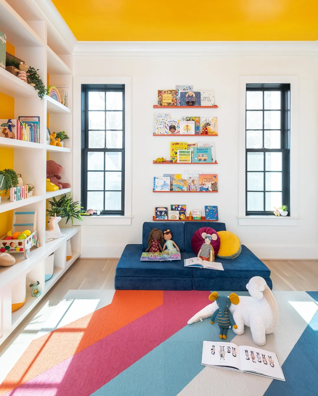 Cozy Reading Nook with Colorful Accents 📚🌟A bright and inviting playroom with a cozy floor sofa, wall-mounted bookshelves, and open cubbies for easy-access toy storage. The bold ceiling color and geometric rug add a playful touch to this beautifully designed space!