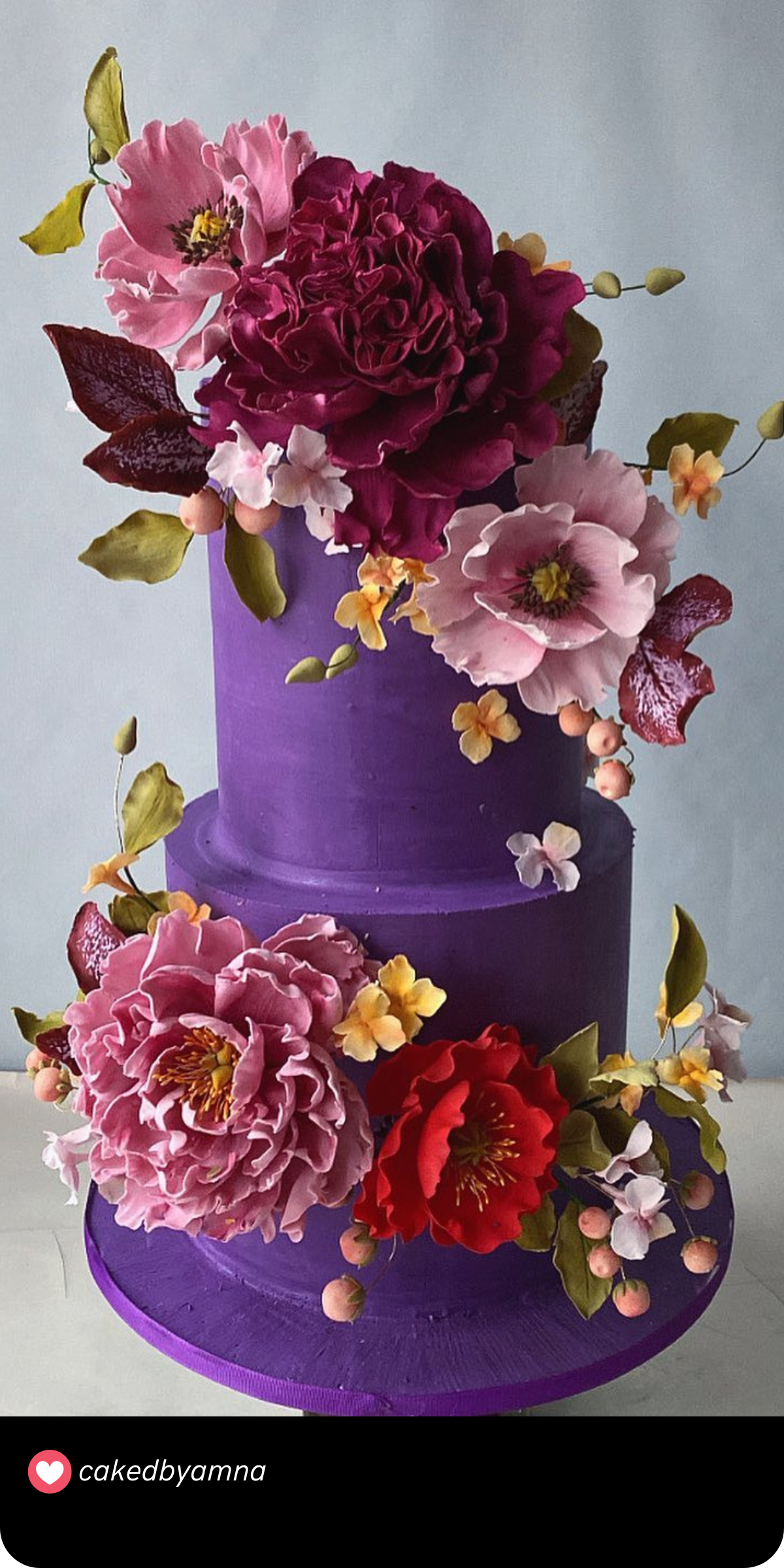 This two-tiered cake in deep purple is beautifully adorned with cascading flowers in shades of pink, burgundy, and peach, creating a lush garden vibe with a vibrant contrast.