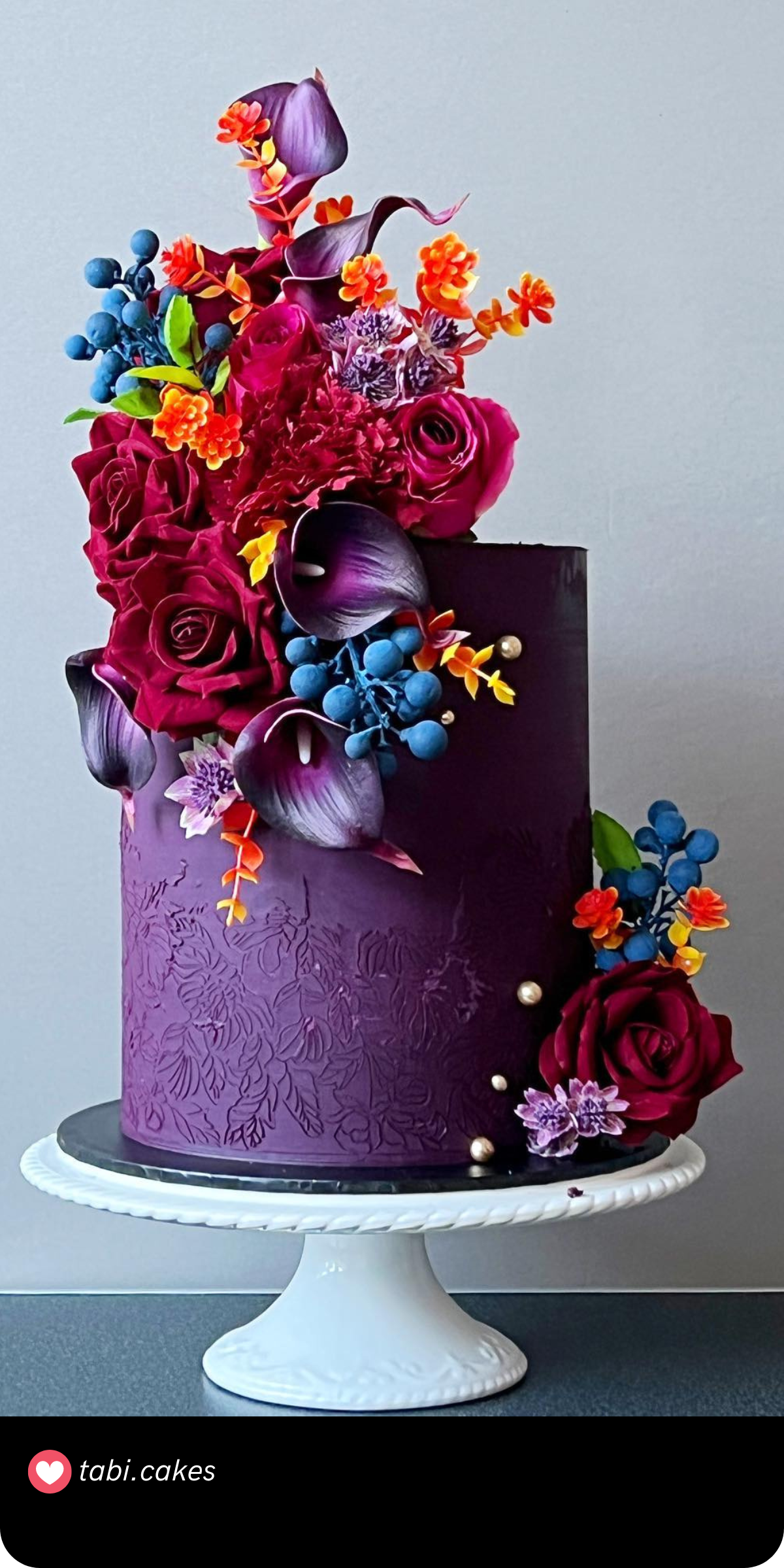 This tall, elegant cake is like a bouquet come to life, featuring deep purple and jewel-toned blooms, accented with playful pops of orange for a chic, artistic feel.