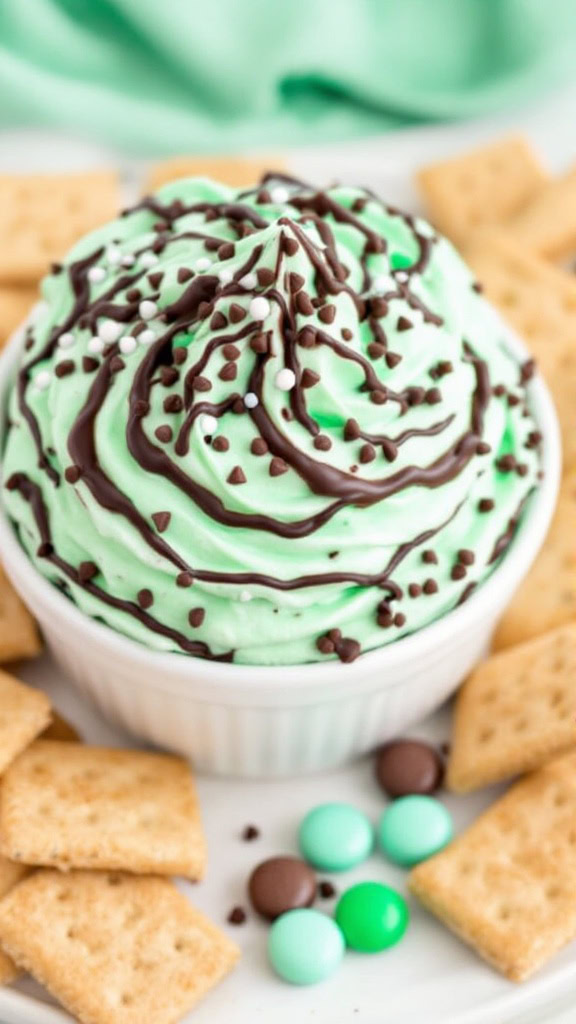 A bowl of mint chocolate cheesecake dip topped with chocolate drizzle and chocolate chips, surrounded by graham crackers and colorful candies.