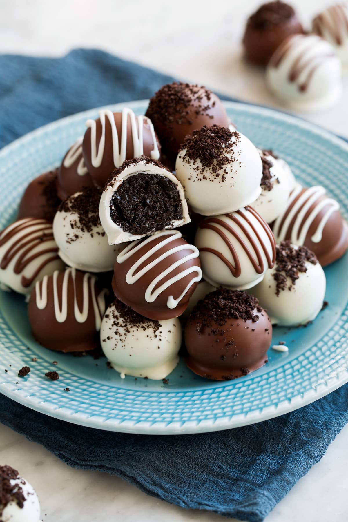 Oreo balls stacked on a turquoise plate set over a blue cloth. Oreo balls are coated in both milk chocolate and white chocolate.